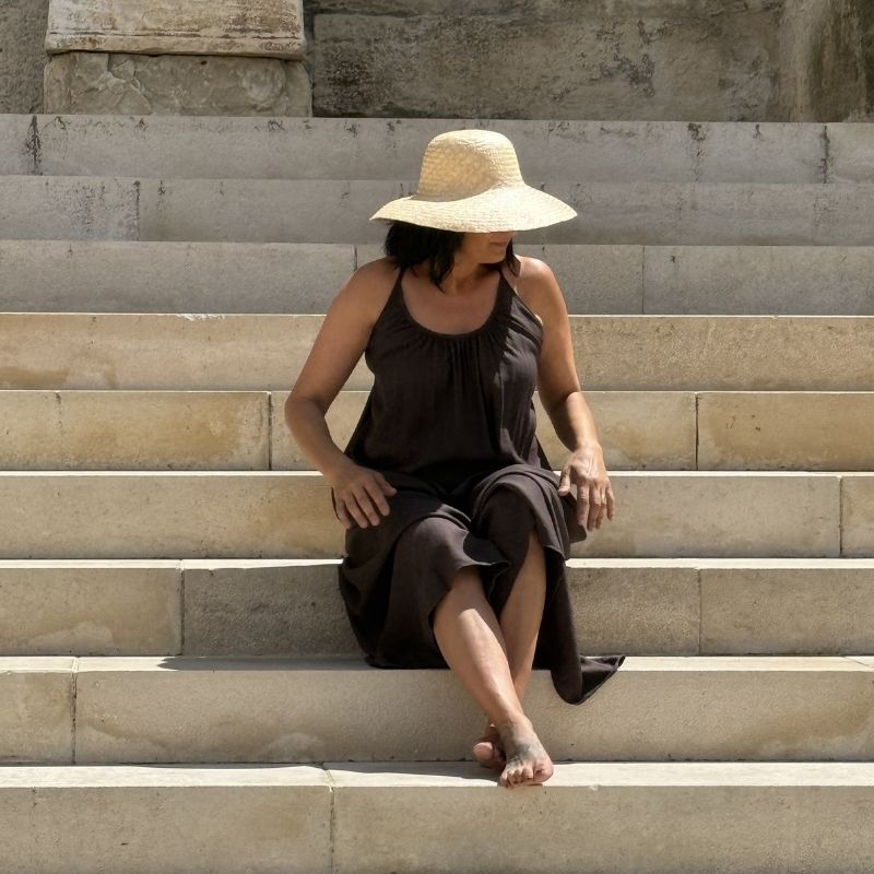 Femme en robe fluide marron avec chapeau sur des marches en pierre, ambiance douce et élégante